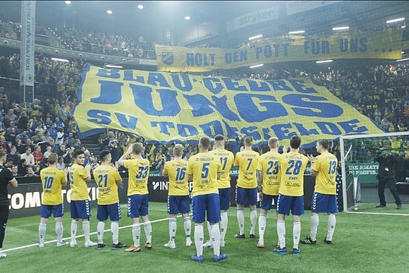 Die Mannschaft des SV Todesfelde lässt sich von ihren Fans feiern. Auf einem großen Banner steht "Blau-Gelbe Jungs SV Todesfelde", auf einem anderen "Holte den Pott für uns".