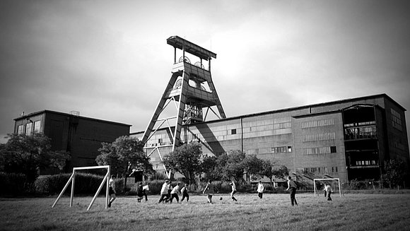 Die schwarzweiß Fotografie zeigt eine Gruppe Kinder beim Fußballspielen auf einer Wiese vor einer Fabrik. 