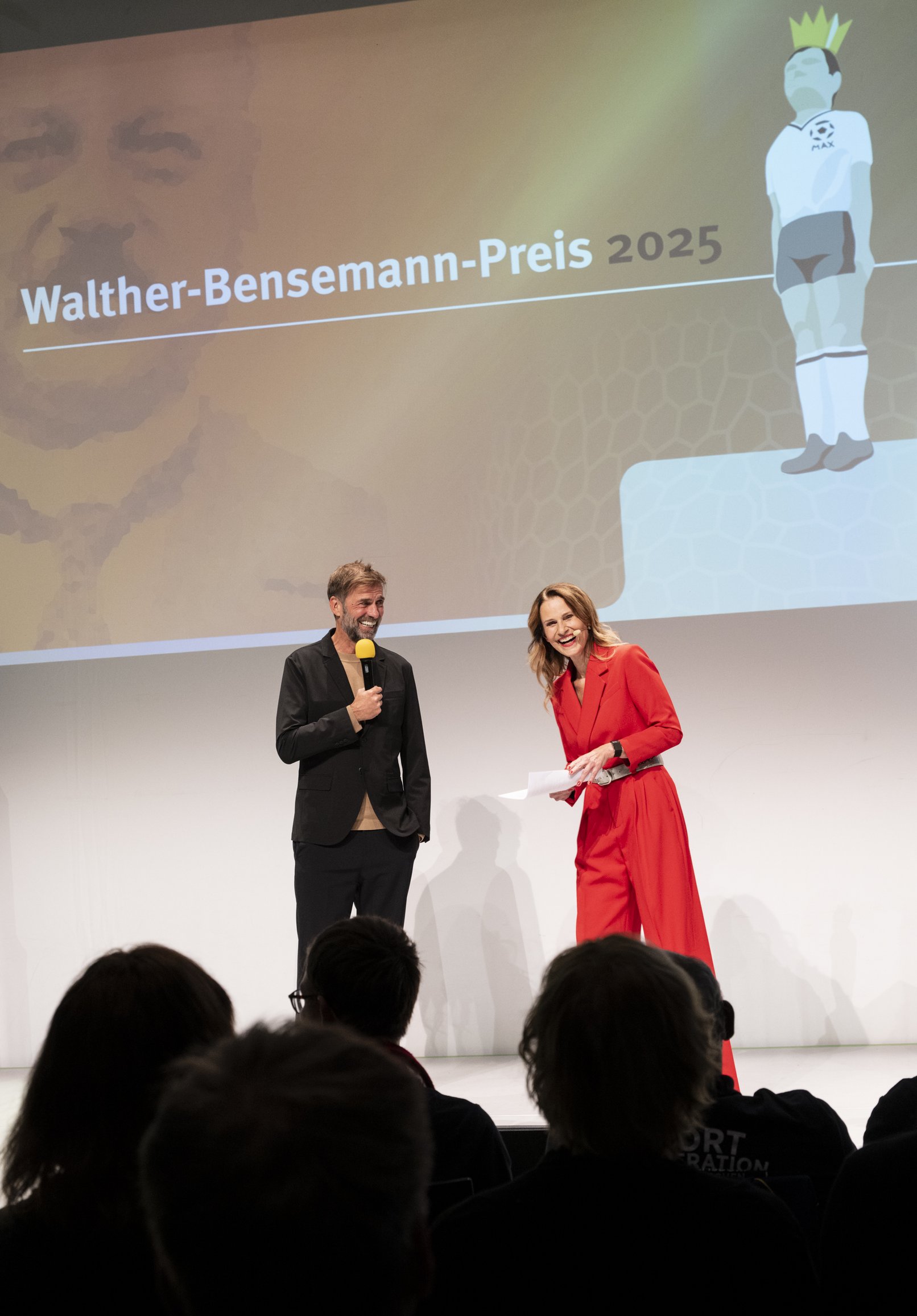 Bensemann-Preisträger Jürgen Klopp gemeinsam mit Moderatorin Kathrin Müller-Hohenstein auf der BÜhne. Im Hintergrund Corporate Design der Gala 2025: organger Hintergrund und Schriftzug Walther-Bensemann-Preis 2025. Daneben die Grafik der Preisfigur MAX mit goldener Krone. 
