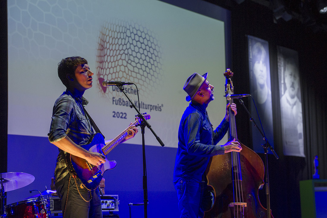 Auf einer blau beleuchteten Bühne stehen zwei Musiker. Link ein Mann mit E-Gitarre, der mit geschlossenen Augen in das Mikrofon vor ihm singt. Rechts neben ihm steht ein Mann mit Hut an einem Kontrabass und singt ebenfalls ins Mikrofon.