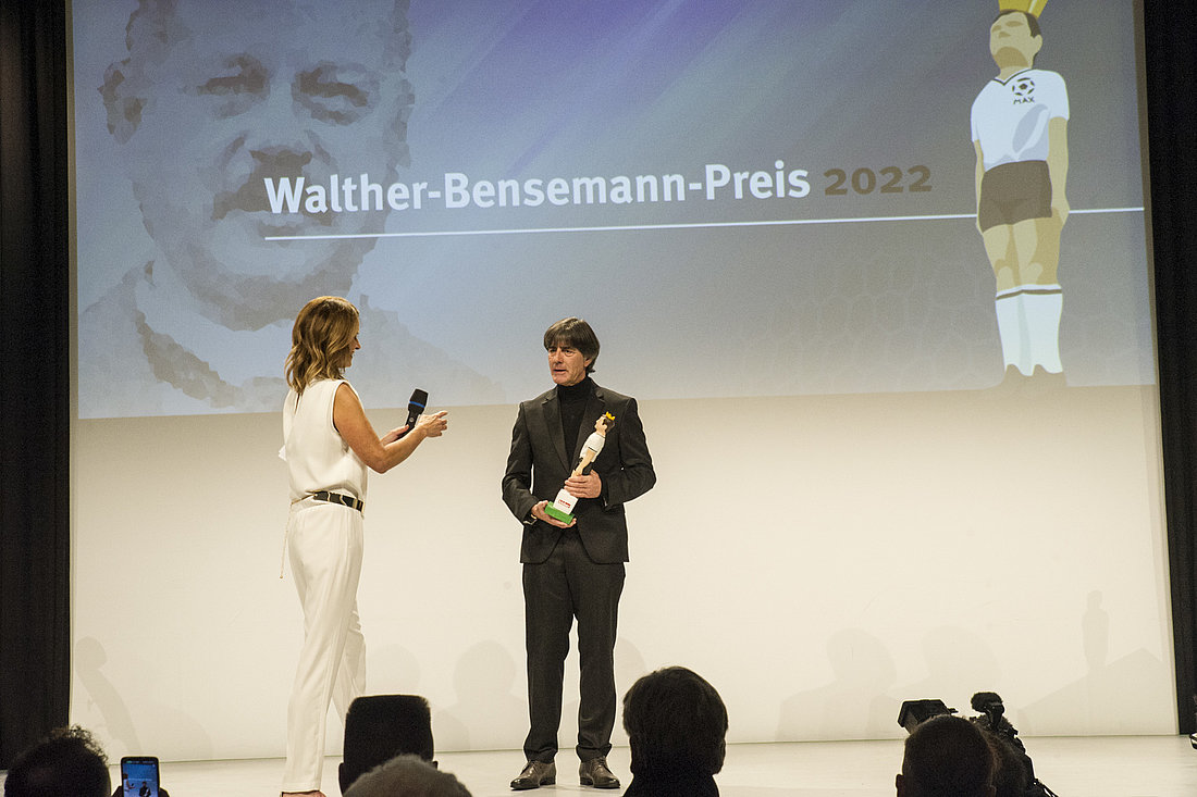 Joachim Löw steht in der Mitte der Bühne neben Katrin Müller-Hohenstein. In den Händen hält er die Siegerfigur.