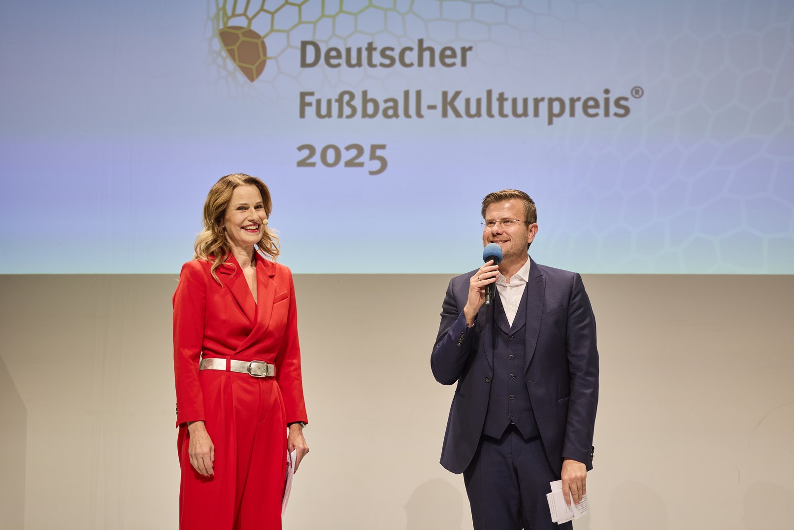 Kathrin Müller-Hohenstien, die druch den Gala-Abend führte und OBM Marcus König begrüßen die Gäste am Beginn der Gala. Im Hintergrund das Logo des Fußball-Kulturpreises 2025, ein als Ball angedeutetes Netz.
