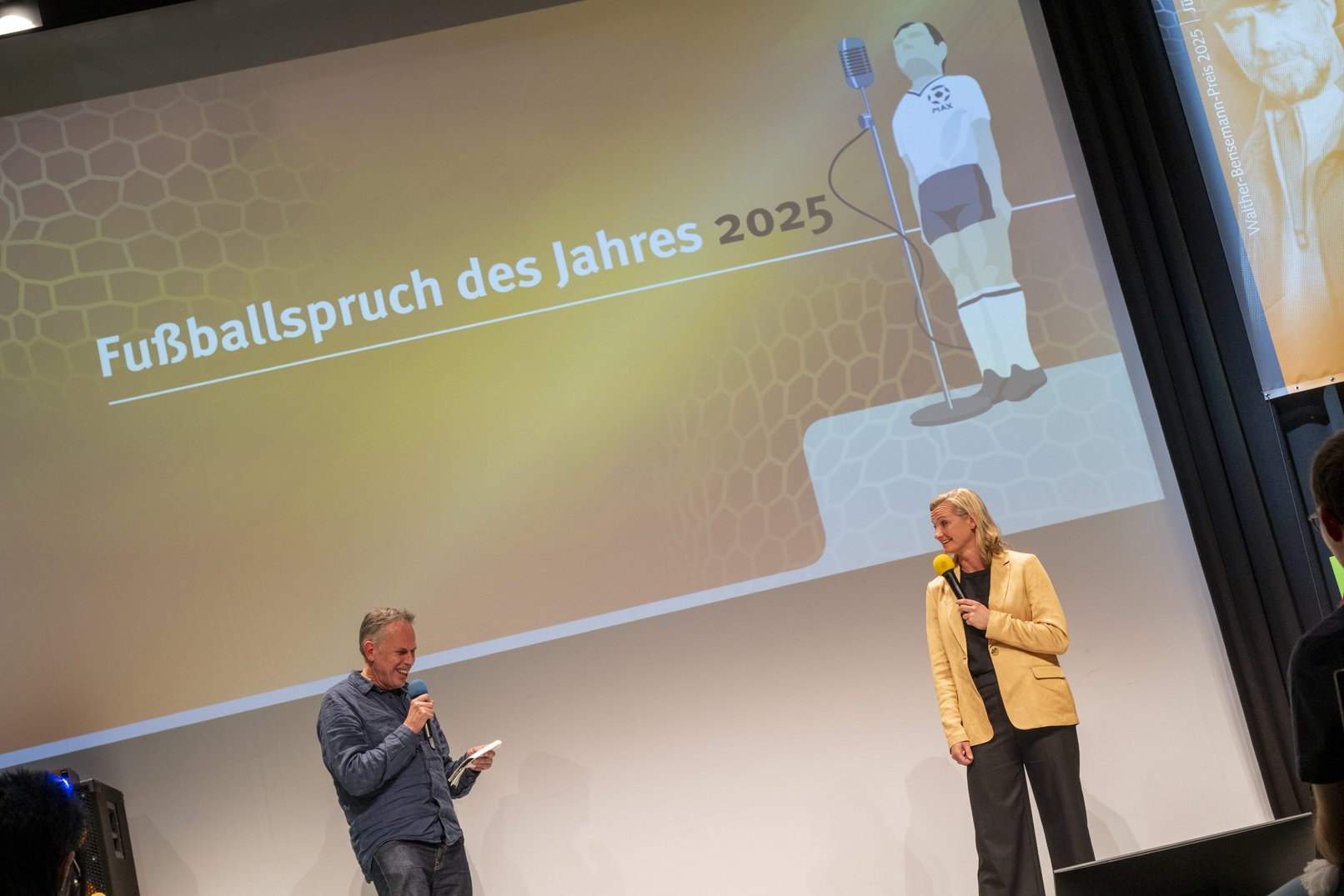 Achim Bogdahn, der die SPruchabstimmung moderierte, gemeinsm mit Alexandra Popp auf der Bühne. Beide lachen. Im Hintergrund das orange Corporate Design der Gala mit dem SChriftzug Fußballspruch des Jahres 2025. Daneben die Grafik der Preisfigur MAX hinter einem Mikro.