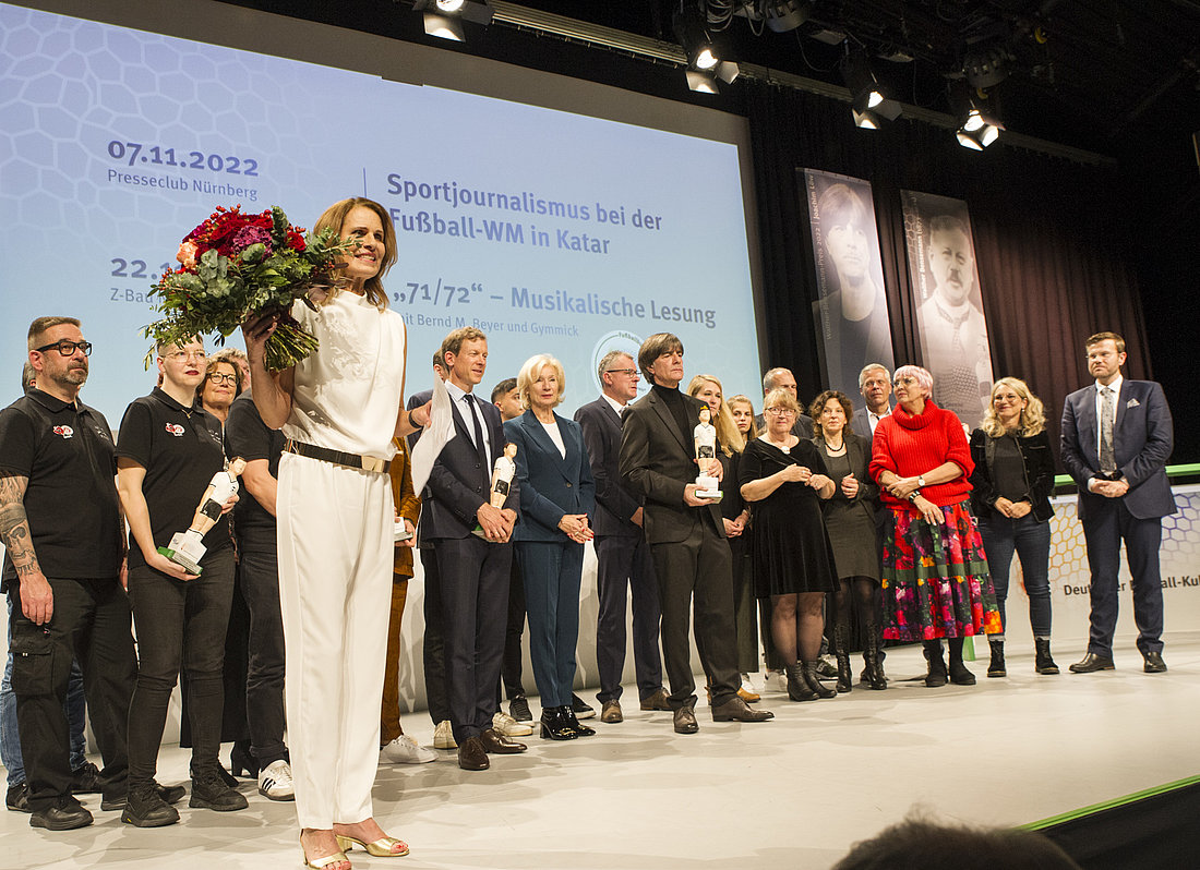 Alle Personen, die bei der Gala einen Bühnenauftritt hatten, kommen zu einem Gruppenbild auf der Bühne zusammen. Die Moderatorin Katrin Müller-Hohenstein steht etwas im Vordergrund und hält einen Blumenstrauß in der Hand. 